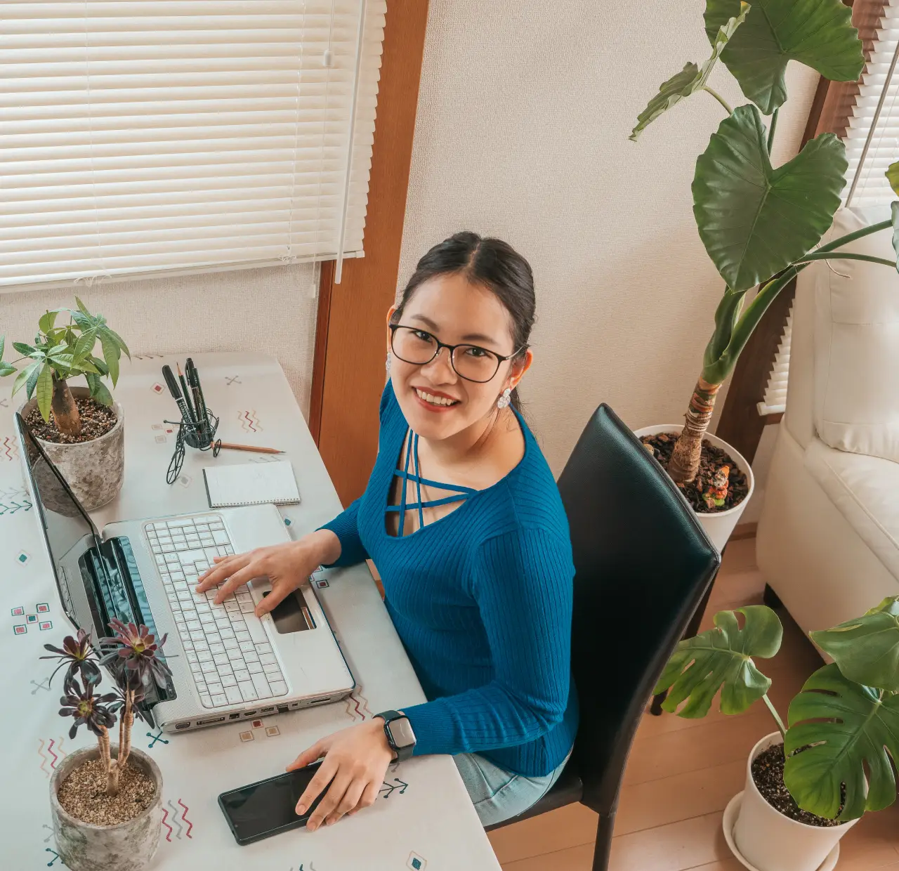 Fille asiatique avec des lunettes, des écouteurs et des plantes travaillant à domicile avec un ordinateur portable et un téléphone portable.